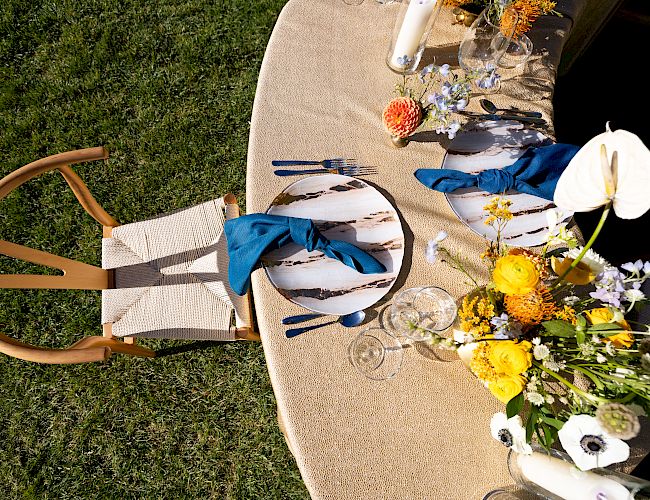 A table set for one with a circular mat, blue napkin, cutlery, and floral arrangements, on grass, next to a wooden chair.