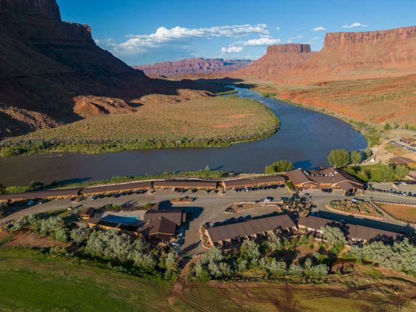 The image shows a river winding through a desert landscape with mesas under a blue sky. In the foreground, there's a rustic building complex.