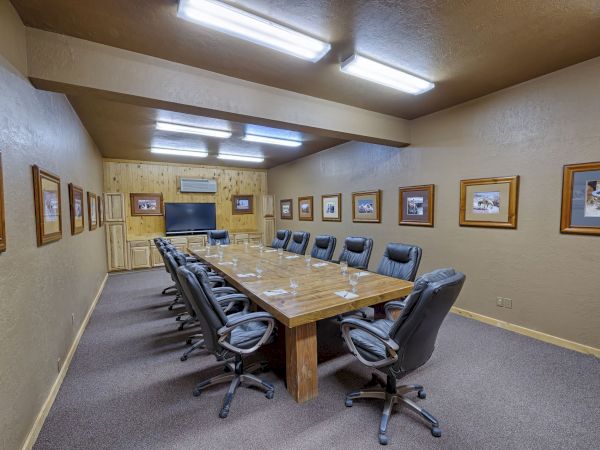 A conference room with a wooden table, black chairs, and framed photos on the walls, under bright ceiling lights.