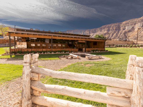 A rustic cabin with a wooden fence, set against a backdrop of rocky cliffs and a cloudy sky, surrounded by green grass and pathways.