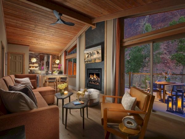 A cozy living room with a fireplace, sofa, armchairs, wood ceiling, and large windows opening to an outdoor patio with a view.