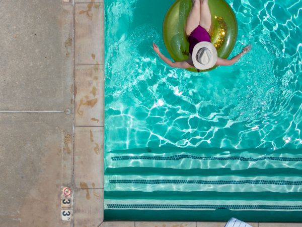 A person in a sun hat floats on a green inner tube in a bright blue pool, relaxing by the edge as sunlight creates shimmering patterns, ending this sentence.