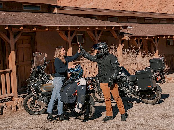 Two people helping a biker with a motorcycle outside a wooden building, desert setting with red rock cliff behind, clear sky.