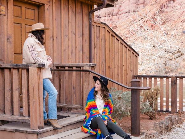Two women wearing sweaters and hats chat in front of their hotel room
