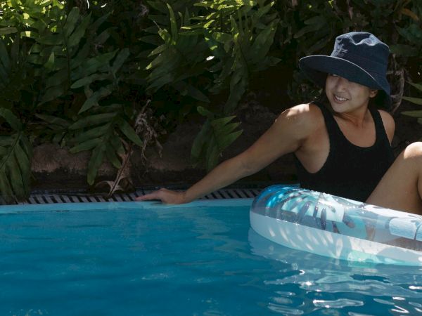 A person in a black swimsuit and hat relaxes on an inflatable in a pool, surrounded by greenery.