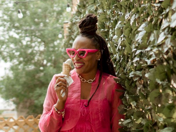 A person in pink sunglasses and a pink outfit enjoys an ice cream cone beside a leafy hedge.