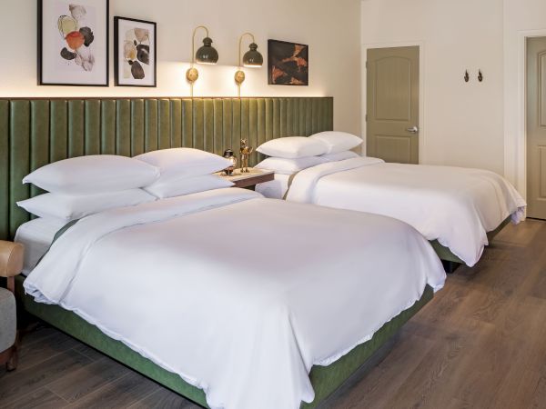 The image shows a hotel room with two neatly made beds, artwork on the wall, a chair, and soft lighting, creating a cozy atmosphere.