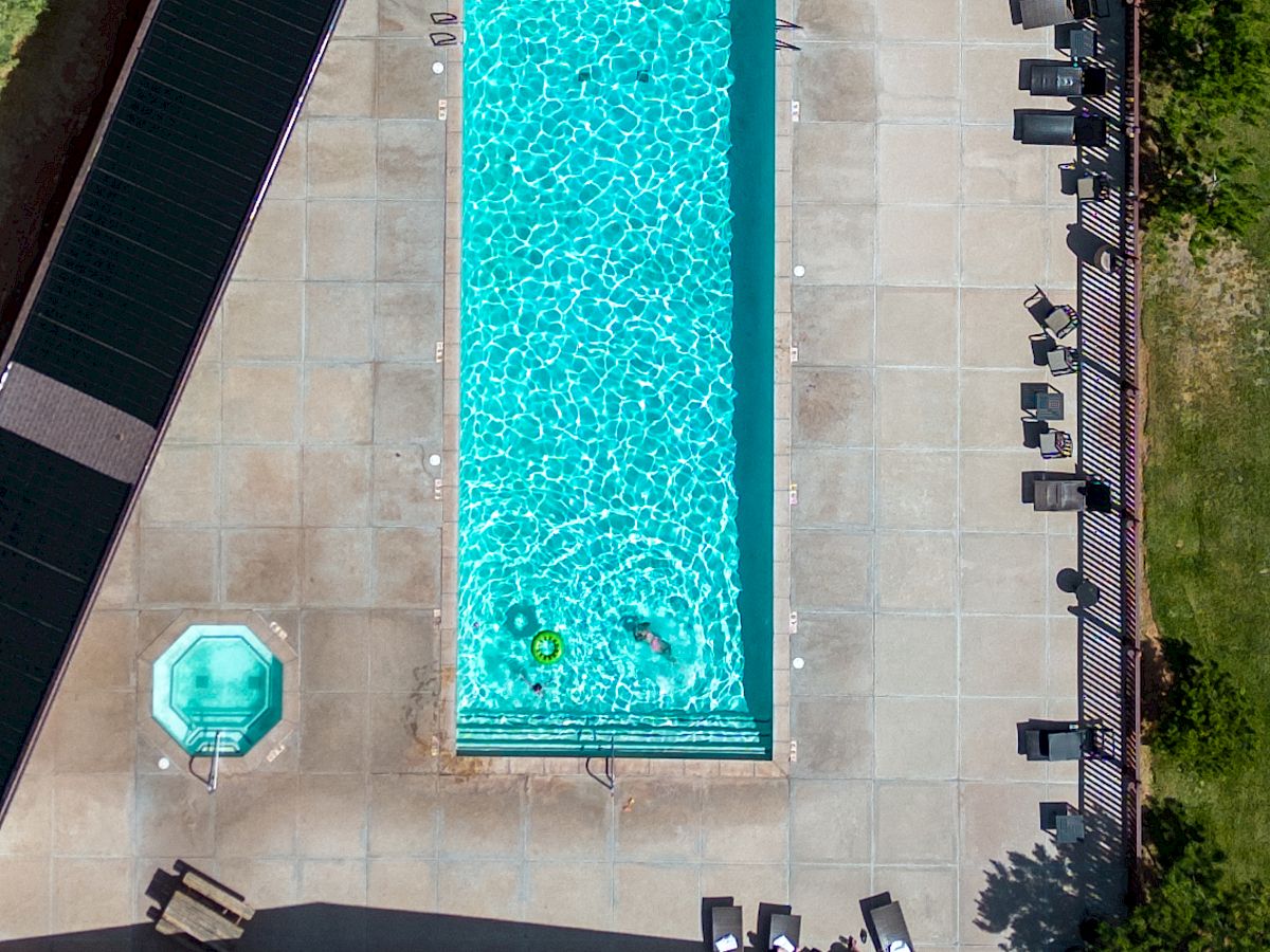 An aerial view of a rectangular in-ground pool with bright blue water, surrounded by a cement patio, lounge chairs, a small round pool, a shed, and yard fencing splitting into a fenced wooded area.