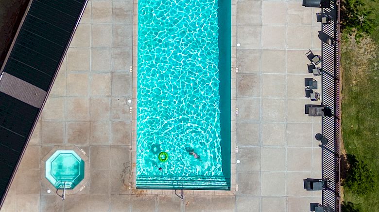 An aerial view of a rectangular in-ground pool with bright blue water, surrounded by a cement patio, lounge chairs, a small round pool, a shed, and yard fencing splitting into a fenced wooded area.