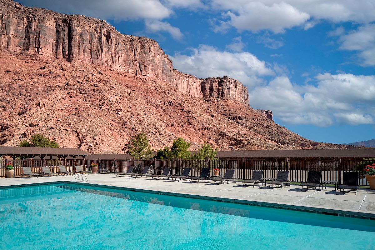 A swimming pool with lounge chairs overlooks a rocky desert landscape under a blue sky with clouds.