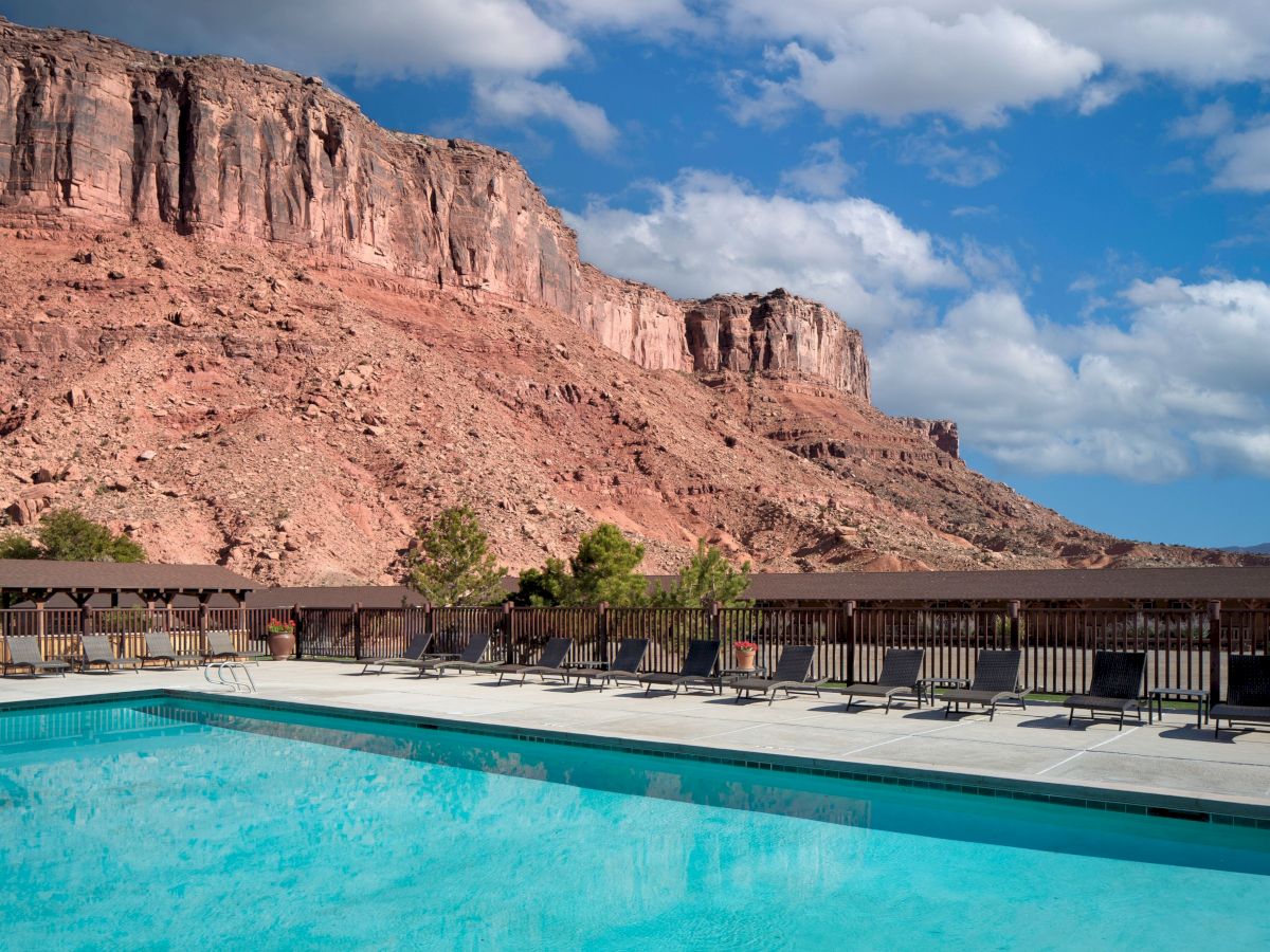A swimming pool with lounge chairs overlooks a rocky desert landscape under a blue sky with clouds.