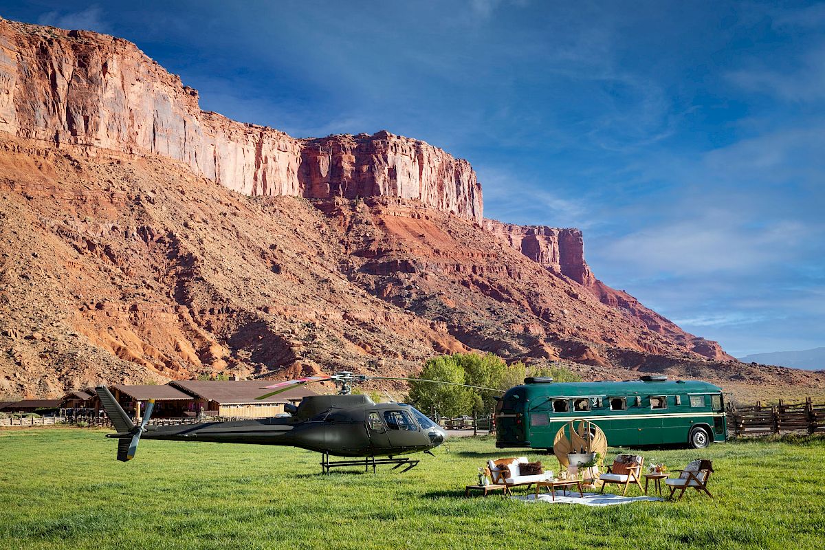 A helicopter, green bus, and outdoor seating set on grass, with dramatic cliffs and a clear blue sky in the background.