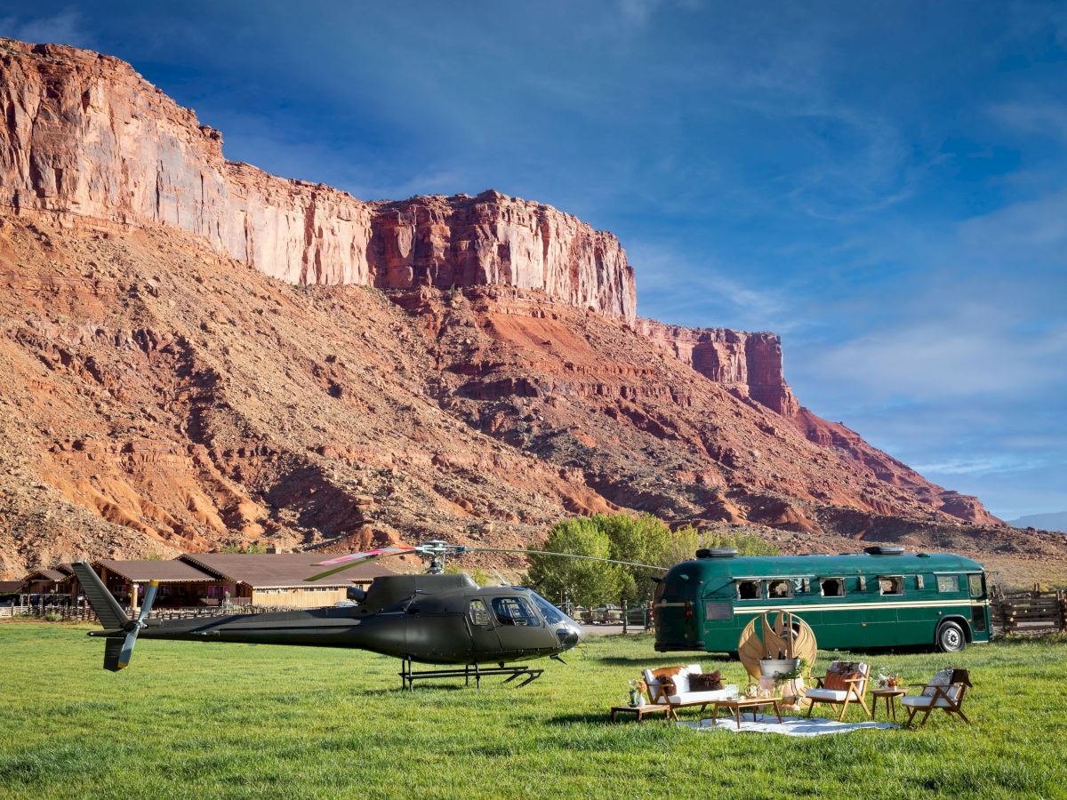 A helicopter, green bus, and outdoor seating set on grass, with dramatic cliffs and a clear blue sky in the background.