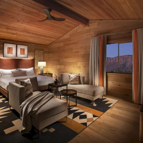 A cozy rustic living room with warm wooden beams, a stone fireplace, sofa, fireplace chair, and framed art, creating inviting cottage charm.