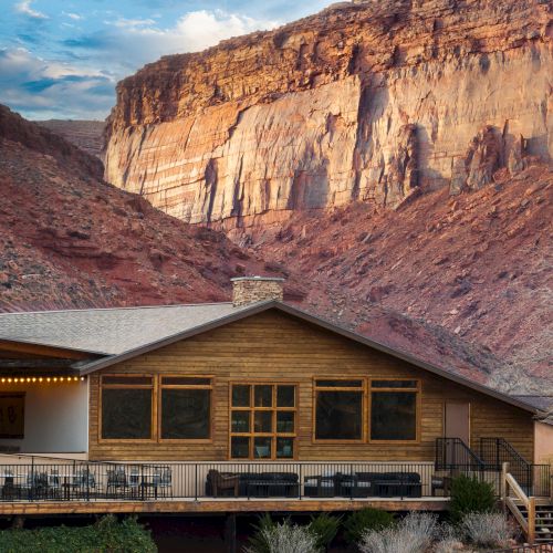 A rustic cabin with a wooden deck sits at the base of dramatic red canyon cliffs, blending rugged landscape with cozy lodging.