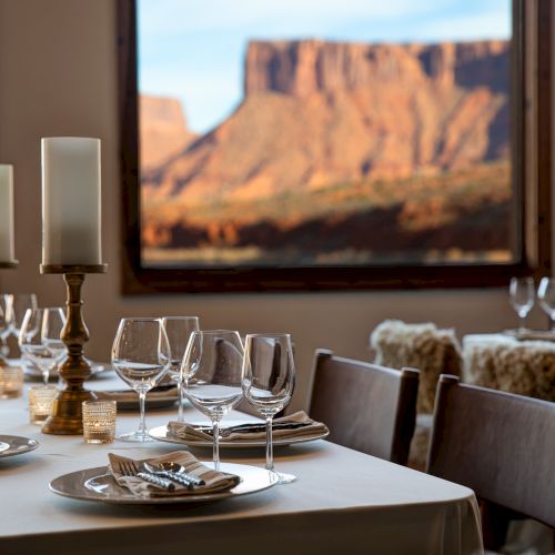 A fine dining table set with white linens, glassware, and candles, overlooking a desert canyon framed by a window, sunset glow.