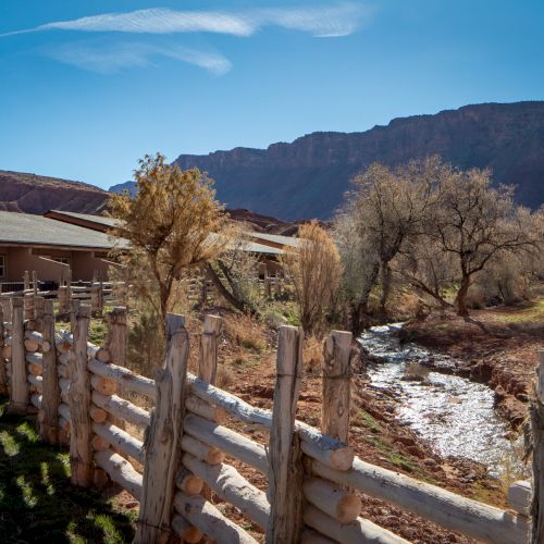 A rustic wooden fence runs along a narrow stream by a stone-walled yard, with desert trees, a building, and red canyon hills in the background.