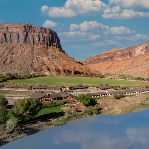 A wide, rocky canyon with layered cliffs, a calm river reflecting the blue sky, and sparse desert vegetation.