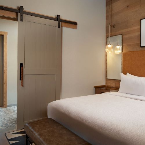 A cozy hotel room with a single bed, warm lighting, a pendant lamp, a wooden desk, and framed pictures on the wall.