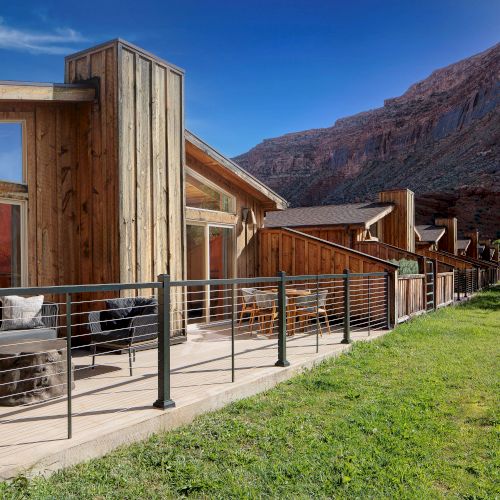 A sunny outdoor patio with rustic wooden furniture and colorful buildings, mountains in the background, and a clear blue sky.