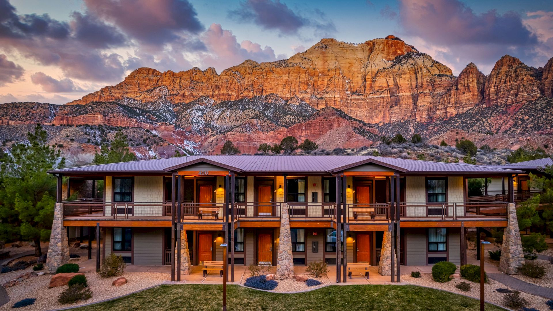 A two-story lodge is set against dramatic, rocky mountains with a colorful sky above, creating a picturesque landscape.