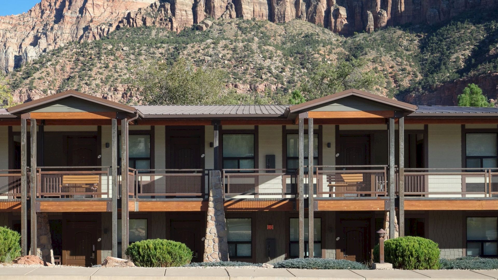 A two-story lodge with wooden accents set against a backdrop of rugged mountains and greenery.