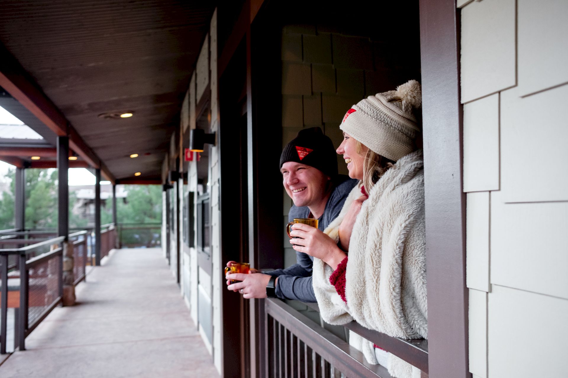 A couple dressed warmly enjoys drinks while looking outside from a covered balcony or porch.