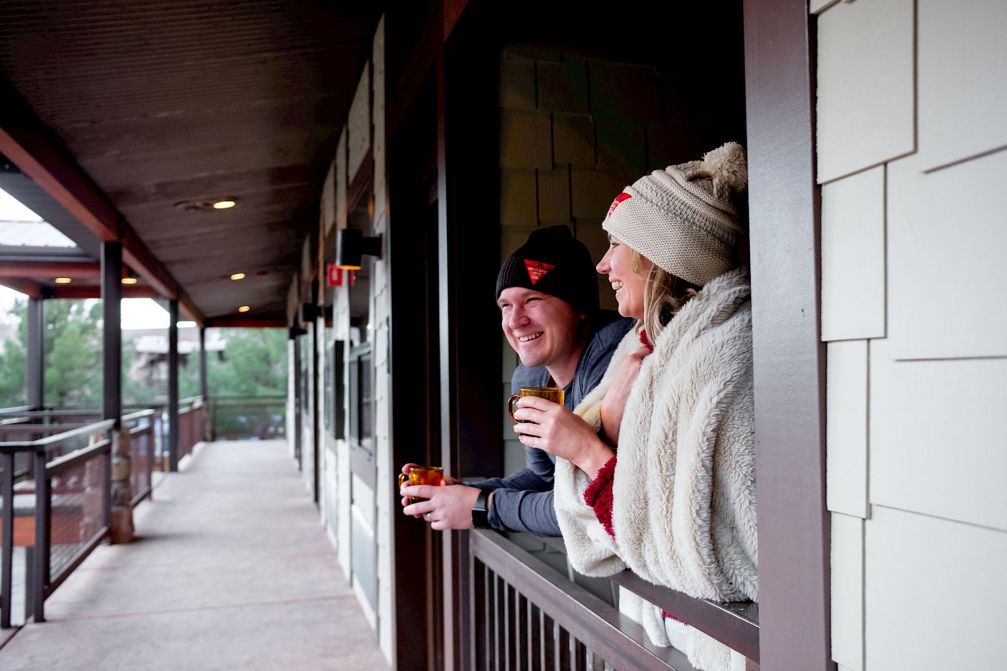A couple dressed warmly enjoys drinks while looking outside from a covered balcony or porch.