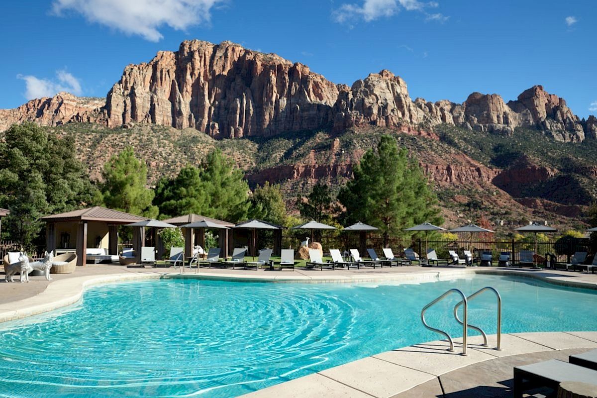 A pool with lounge chairs and cabanas surrounded by lush trees and majestic mountains under a clear blue sky.