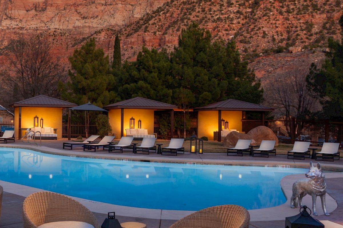 A serene pool area with wicker chairs and lanterns is set against a backdrop of towering red rocks and softly lit cabanas at dusk.
