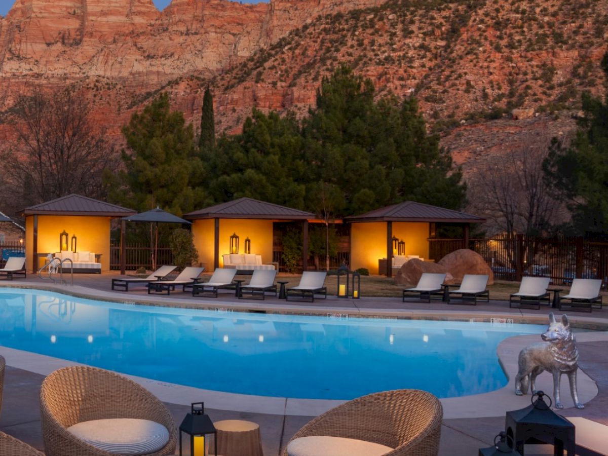 A serene pool area with wicker chairs and lanterns is set against a backdrop of towering red rocks and softly lit cabanas at dusk.