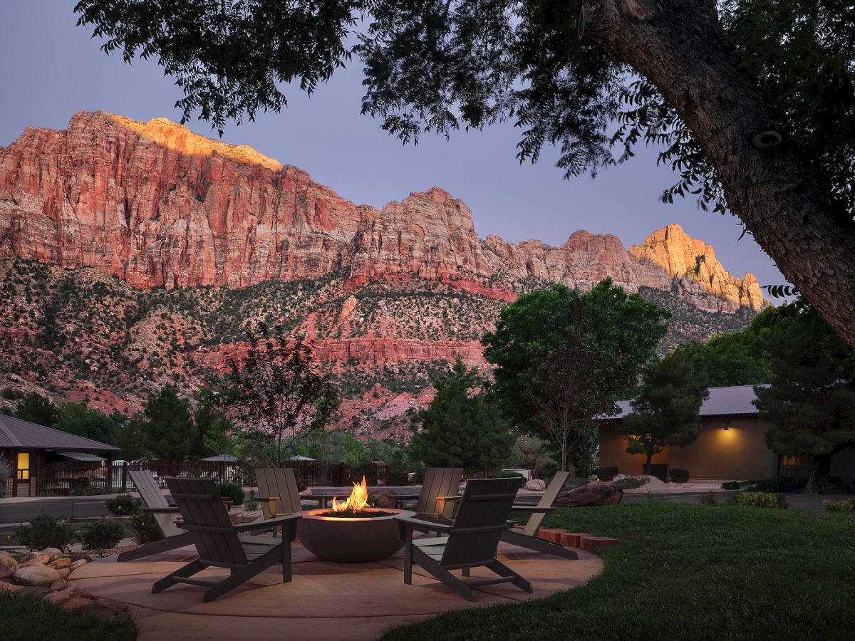 A cozy outdoor seating area with a fire pit surrounded by chairs, set against a backdrop of red glowing canyon cliffs at dusk.