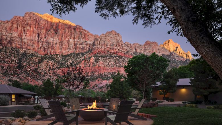 A cozy outdoor seating area with a fire pit surrounded by chairs, set against a backdrop of red glowing canyon cliffs at dusk.