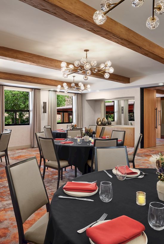 A stylish restaurant dining room with round tables, black tablecloths, red napkins, glassware, candles, and warm lighting.