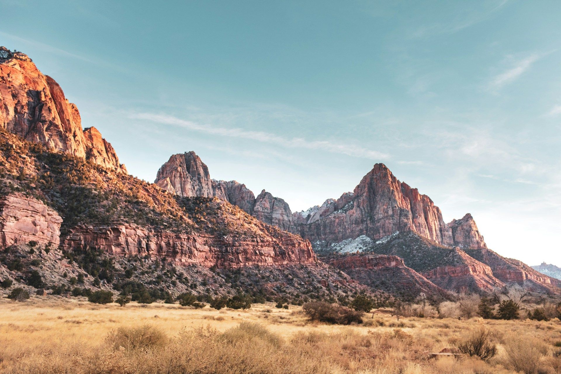 The image shows a stunning view of a mountainous landscape with red and brown cliffs and a clear blue sky, surrounded by dry grass.