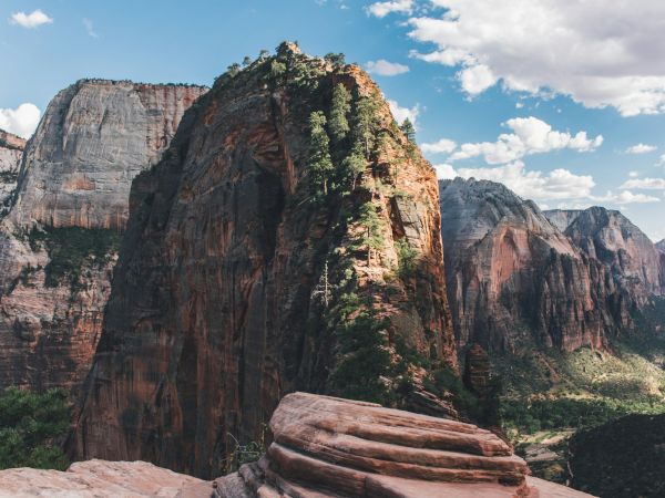 A stunning view of a towering rock formation in a canyon, under a blue sky with clouds, surrounded by lush greenery and rugged cliffs.