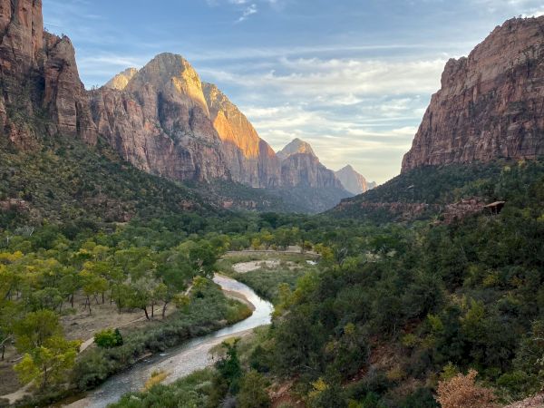 A scenic view of a winding river flanked by lush greenery and towering mountains under a partly cloudy sky.