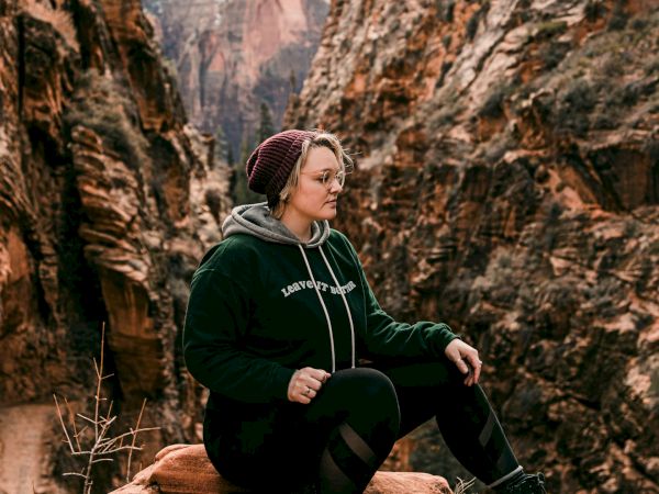 A person sits on a rock in a canyon, wearing a hoodie and beanie, with a mountainous landscape in the background.