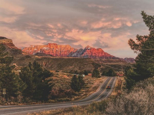 A scenic road leads to majestic mountains under a dramatic sky, with vibrant sunset hues highlighting the landscape.