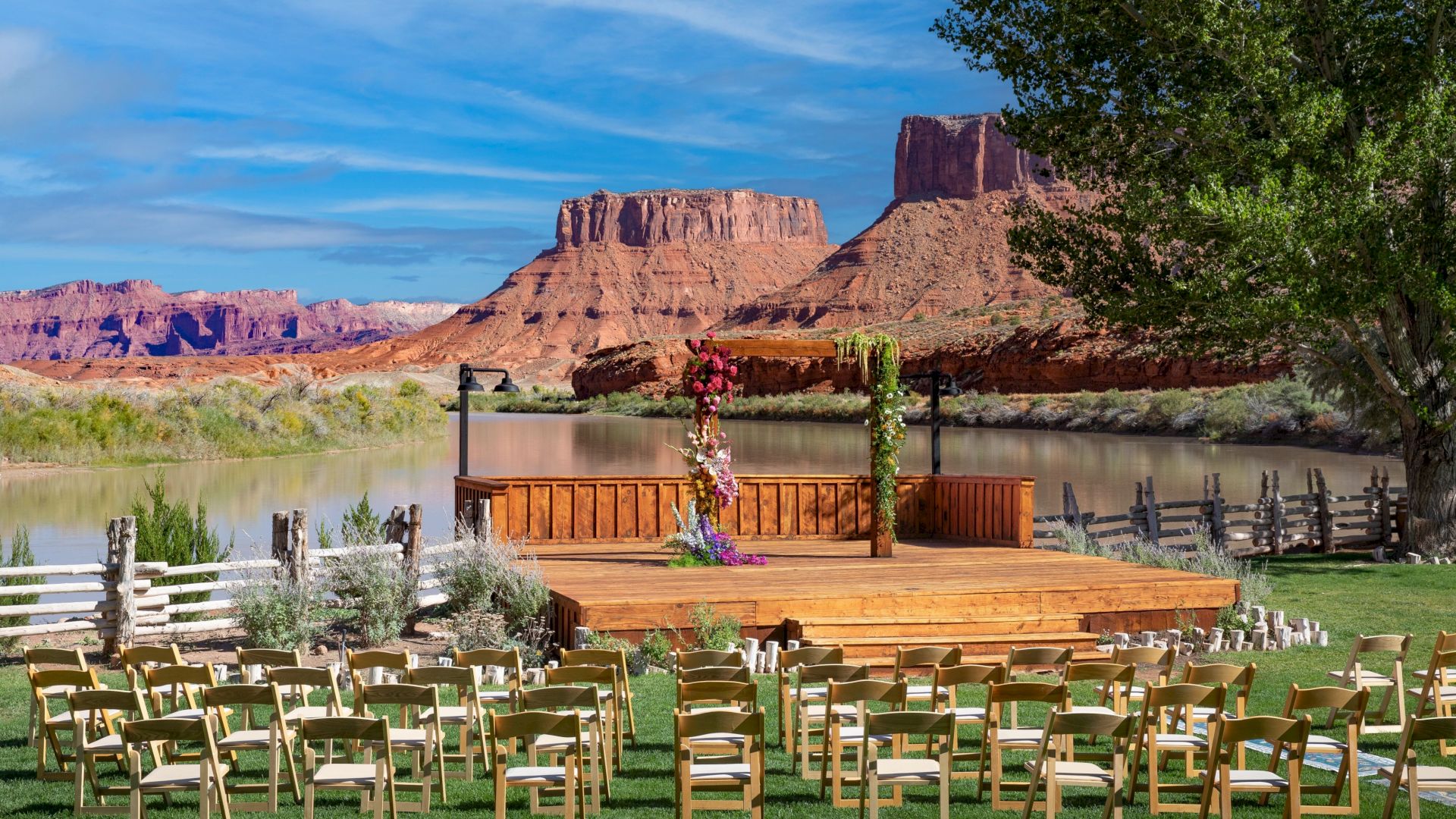 An outdoor wedding setup with chairs facing a decorated wooden platform, set against a backdrop of red rock formations and a river.