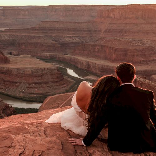 A couple sits on a rocky overlook, watching a river winding through a canyon at sunset, creating a romantic and serene atmosphere.