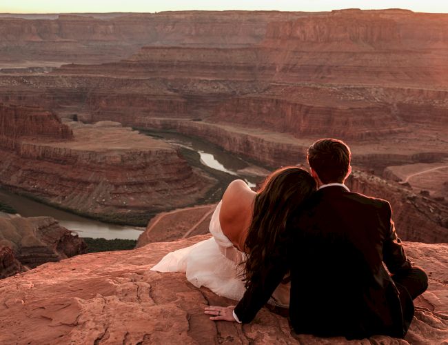 A couple sits on a rocky overlook, watching a river winding through a canyon at sunset, creating a romantic and serene atmosphere.