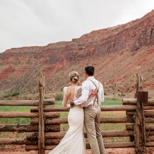 A couple in wedding attire stands by a wooden fence, overlooking a scenic landscape with red rock formations and a cloudy sky.