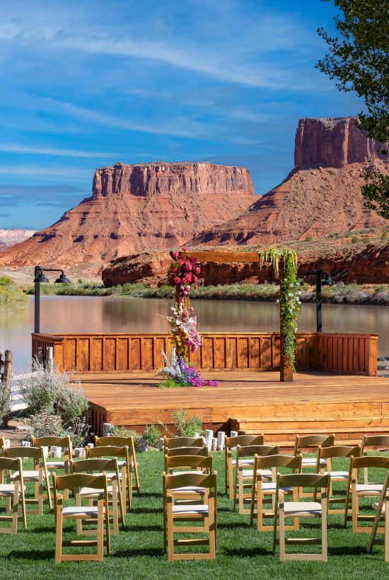 An outdoor wedding setup with chairs on a lawn, a decorated wooden stage, and a scenic background of red rock formations near a river.