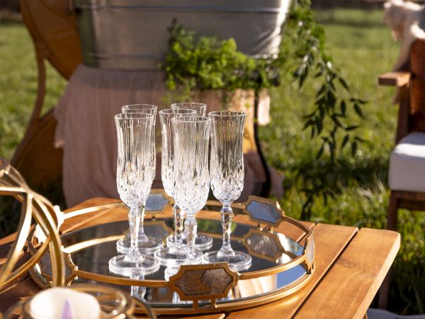 A set of elegant crystal champagne flutes on a reflective tray, placed on a wooden table outdoors with greenery in the background.