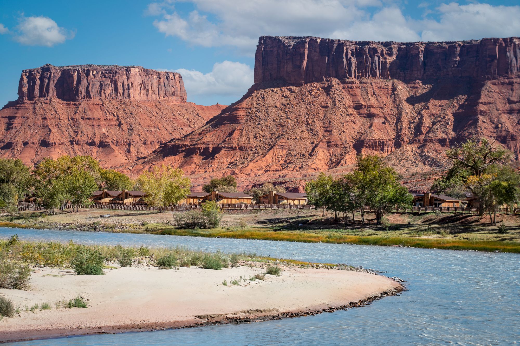 A river flows through a desert landscape with red rock cliffs and scattered trees under a blue sky with clouds.