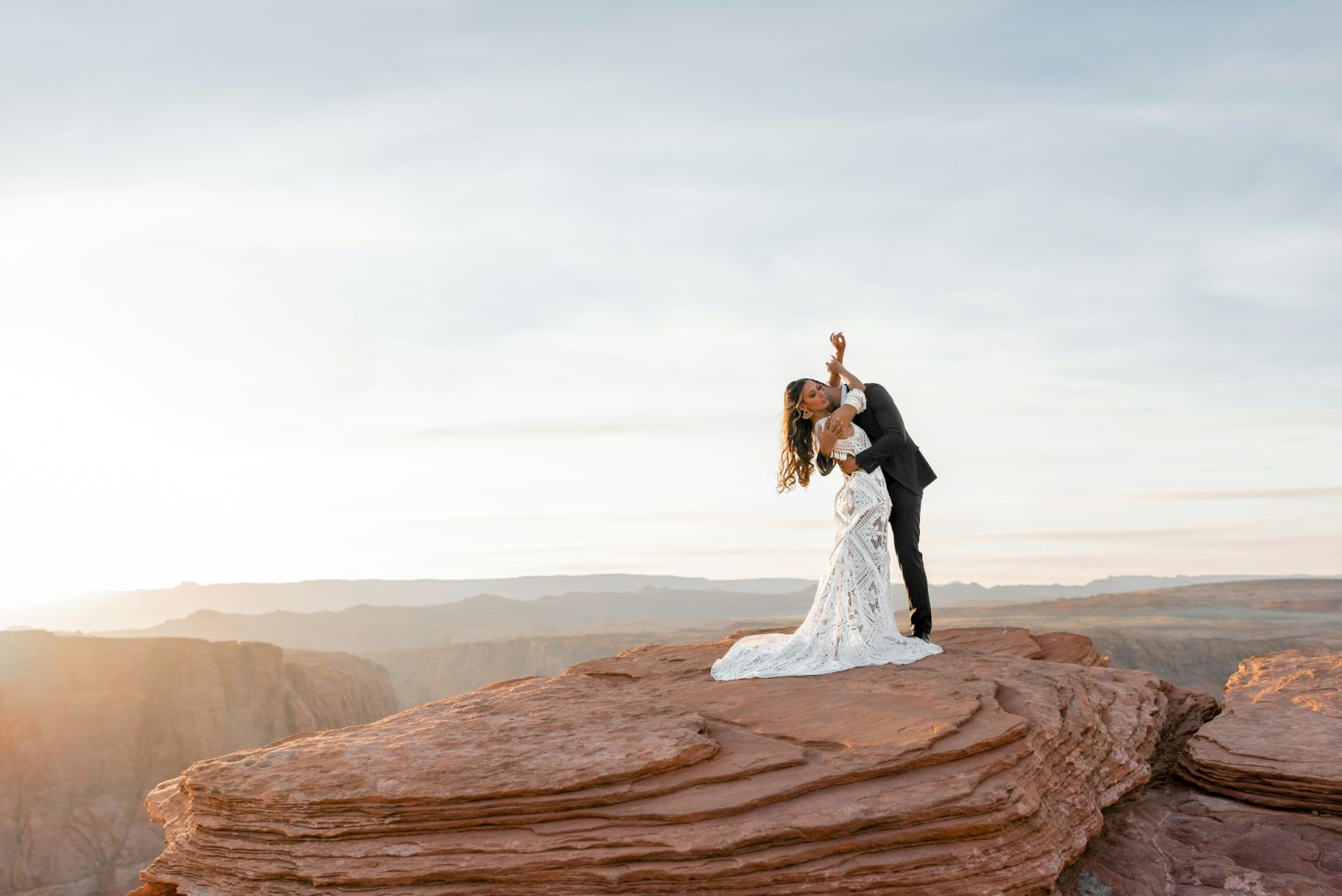 A couple in wedding attire poses on a rocky cliff at sunset, with a breathtaking landscape in the background.