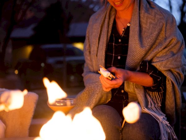 A person wrapped in a blanket is roasting marshmallows over an outdoor fire.