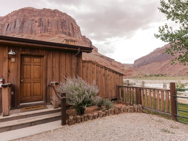 A rustic wooden cabin sits in a desert landscape with red rock formations and a cloudy sky in the background.