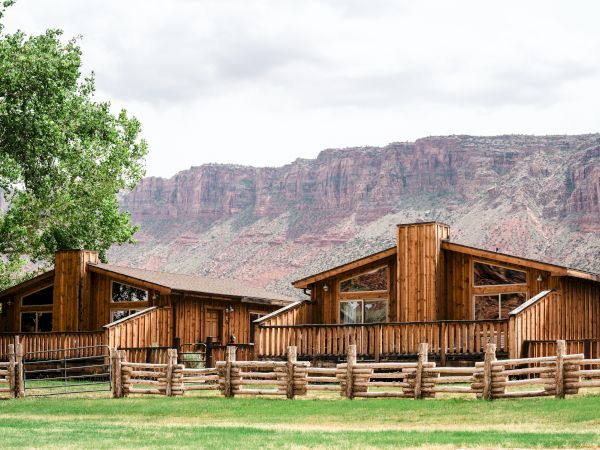 Two rustic wooden cabins are set against a backdrop of rocky cliffs and green grass, with a tree nearby and a wooden fence in the foreground.